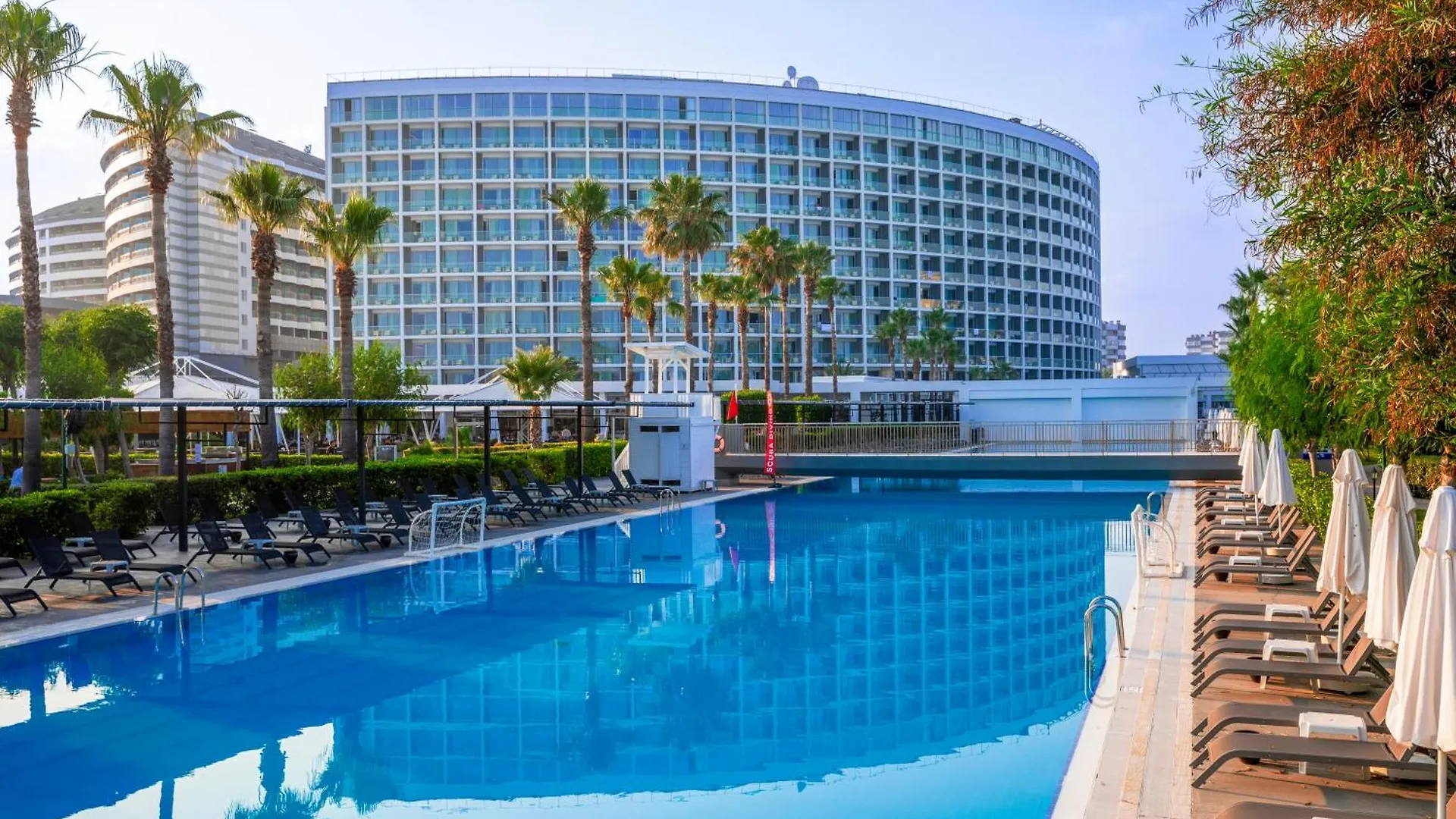 Crystal Centro Pearl Collection Hotel Antalya Exterior photo This is a bright, sunny photo showcasing a luxurious outdoor setting, likely at a high-end hotel or resort.

The focal point in the background is a very large, modern, curved high-rise building with a glass facade, reflecting the bright sky. Palm tre